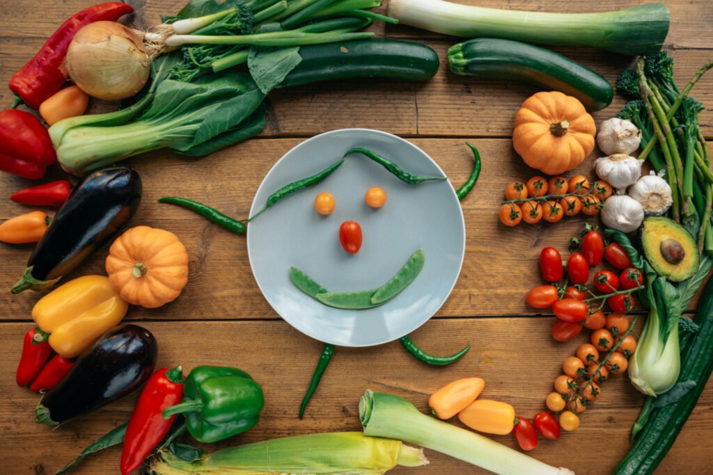 Fresh vegetables arranged creatively around a smiling plate on wooden table.